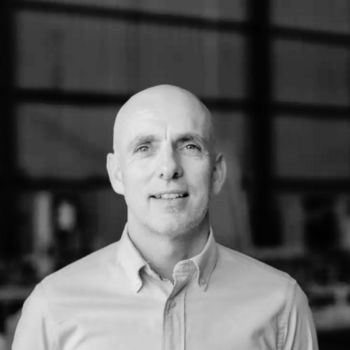 Black-and-white portrait of a bald man in a collared shirt, standing indoors.